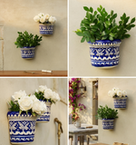 Two handcrafted blue pottery wall hanging pots with blue and white patterns mounted on a rustic stone wall, one holding white roses and the other green foliage.