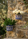 Two handcrafted blue pottery wall hanging pots with blue and white patterns mounted on a rustic stone wall, one holding white roses and the other green foliage.